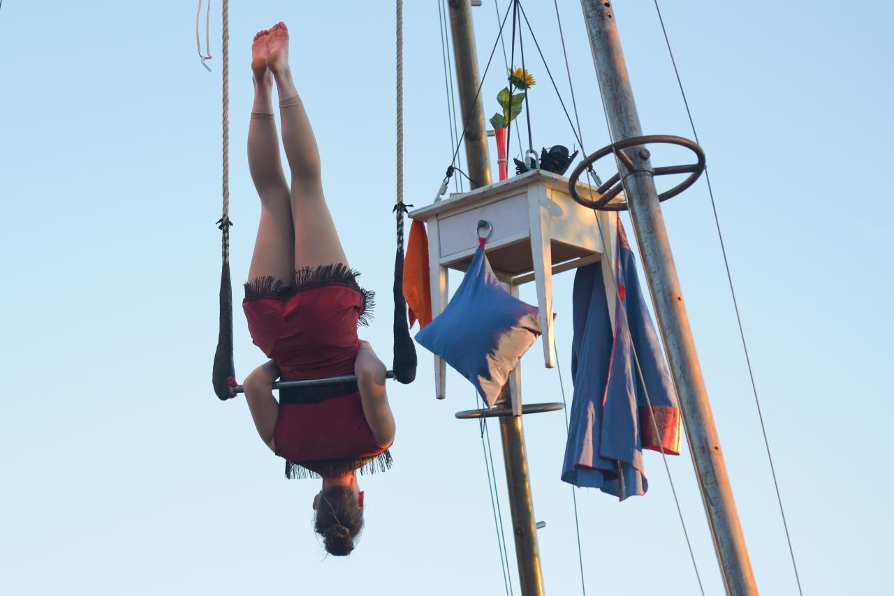 Jana Korb in der Rolle der Trapezkünstlerin schwingt an den Ellbogen um ihr Trapez. Sie hängt am Trapezgerüst neben einem Tischchen mit Kissen und Kleidung vor blauem Himmel.
