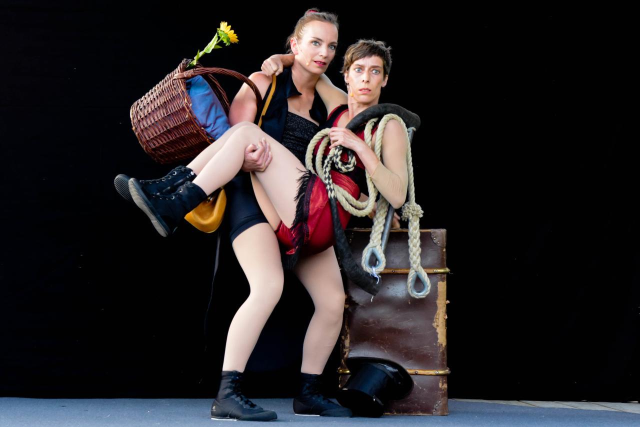 Die Impresaria hält die Trapezkünstlerin im Arm, mit all ihrem Gepäck, einem Korb, einer Tasche und dem Trapez. Beide Performerinnen sind weiße cis Frauen.