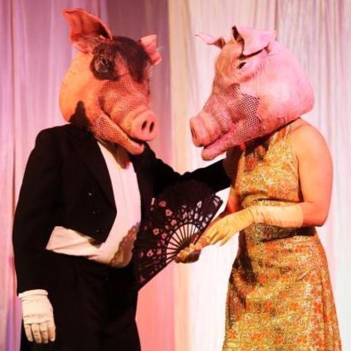 Schweinekapitalist*innen in Abendgarderobe: zwei rosa Hausschweine (Stelzenläufer*innen) in Frack und goldenem Abendkleid und rosa Schweinemasken.