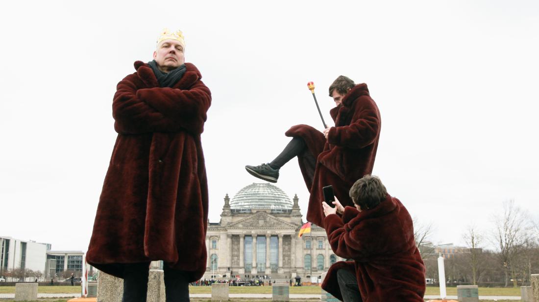 Drei Personen in weinroten Mänteln stehen vor dem Reichstagsgebäude. Einer trägt eine Krone, ein anderer tritt perspektivisch die Reichstagskuppel ein, während die dritte Person ein Foto davon macht.